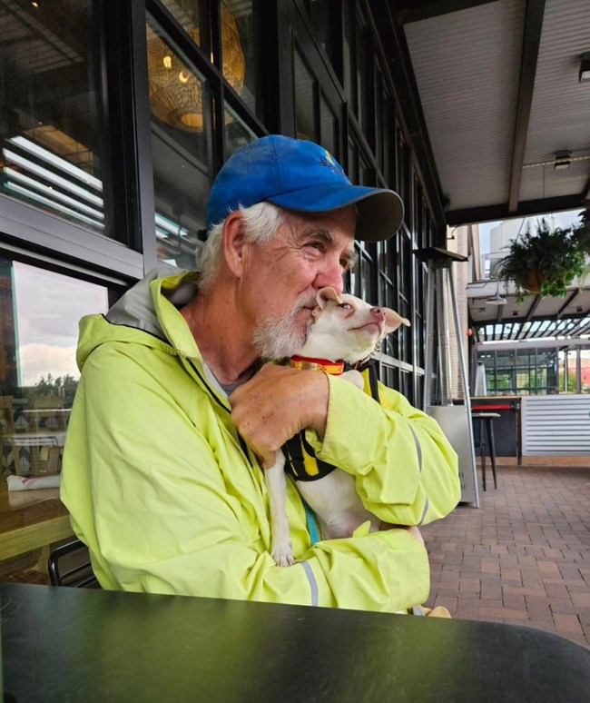 A man sitting at an outdoor table holds a small dog in his arms while nuzzling its head.