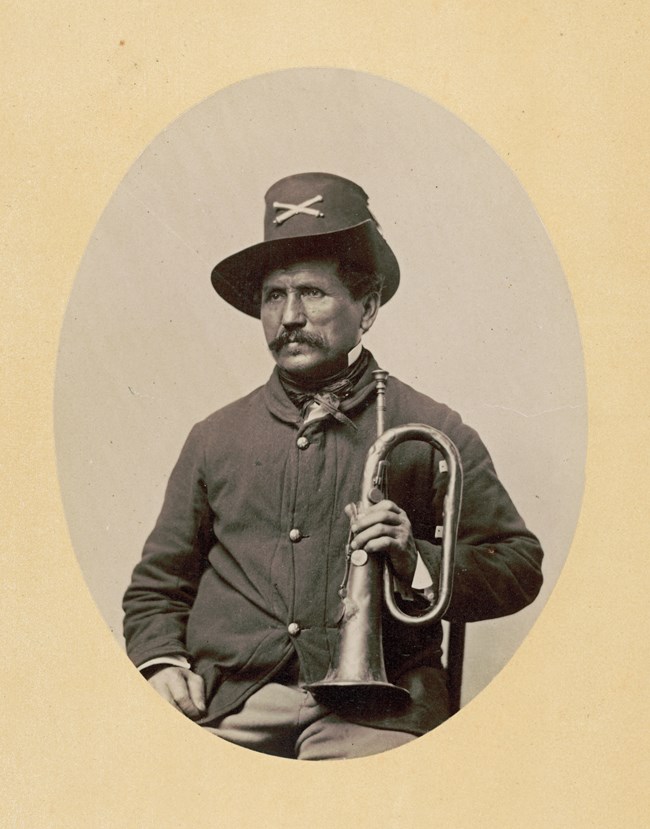 A middle aged man poses for the camera in a 19th century US military uniform standing a bugle on his lap.