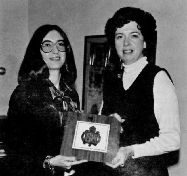 Cecilia Matic and Lorraine Mintzmyer in street clothes pose as they both hold onto a square plaque.