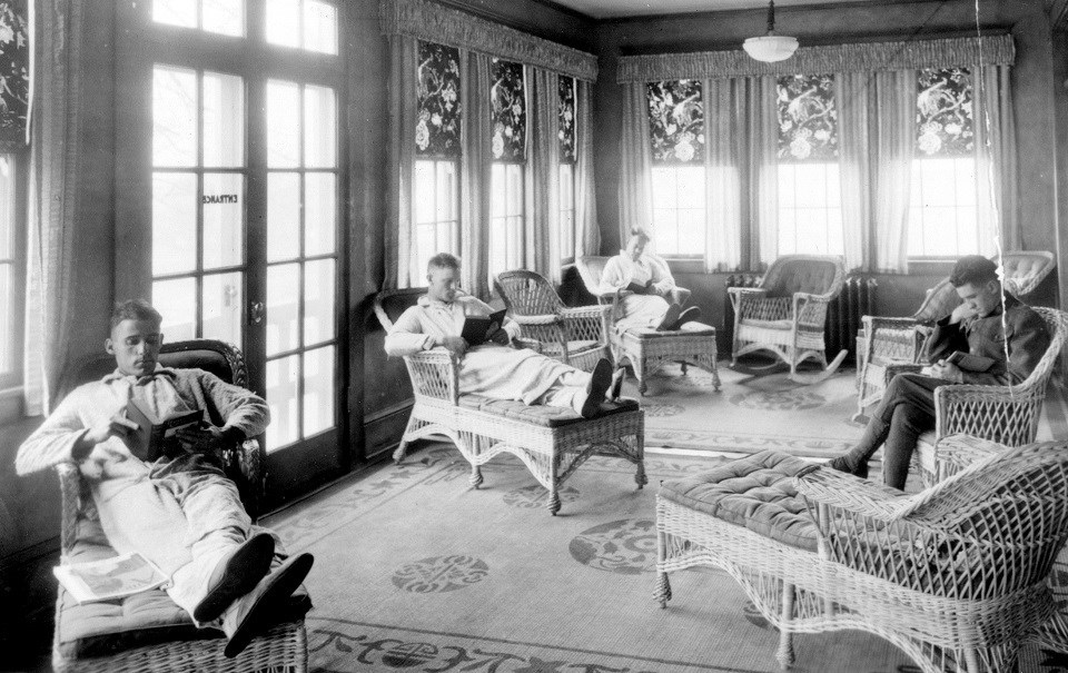 A black and white photo of men wearing robes sitting in a room with large windows. The furniture is wicker and the men have their feet up on footstools while they read books.