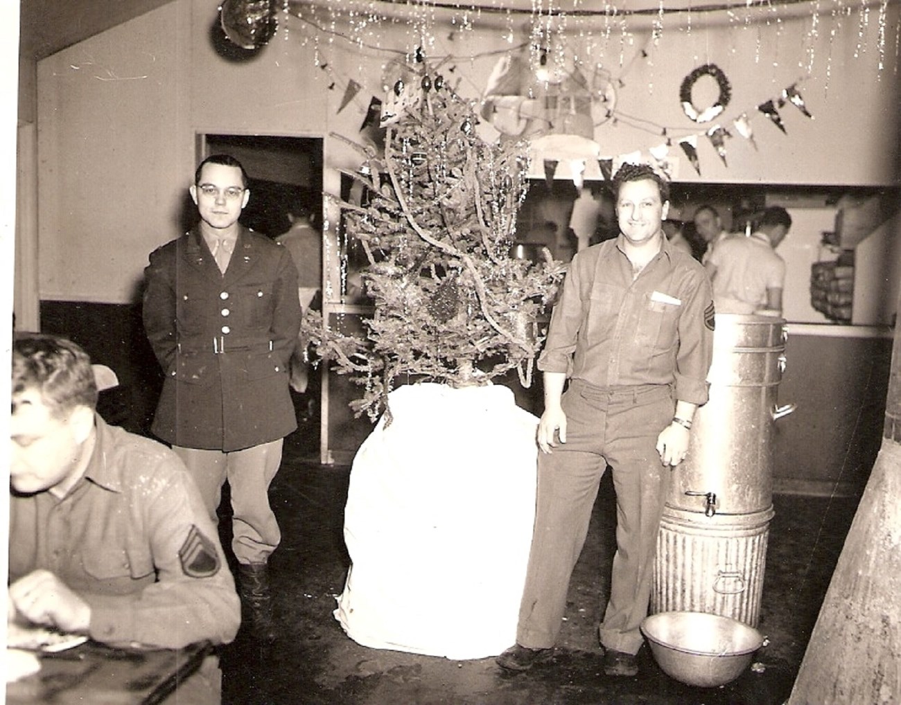 Black and white photo of two men standing beside a small, decorated Christmas tree in the mess hall.