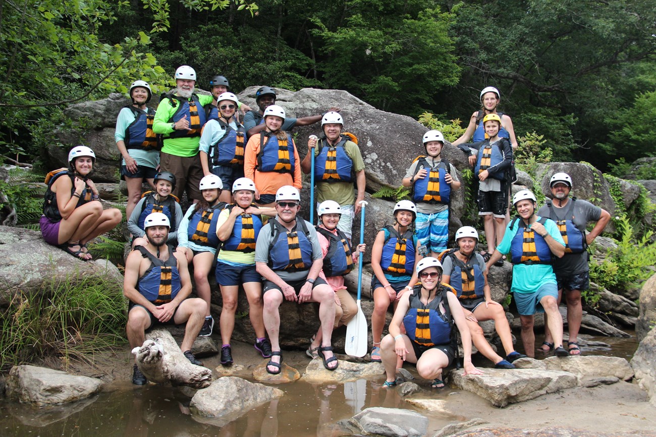 Large group poses for group photo wearing personal flotation devices (life jackets) and helmets. Two people hold paddles. Front row sits on large rocks along shallow creek; others stand behind