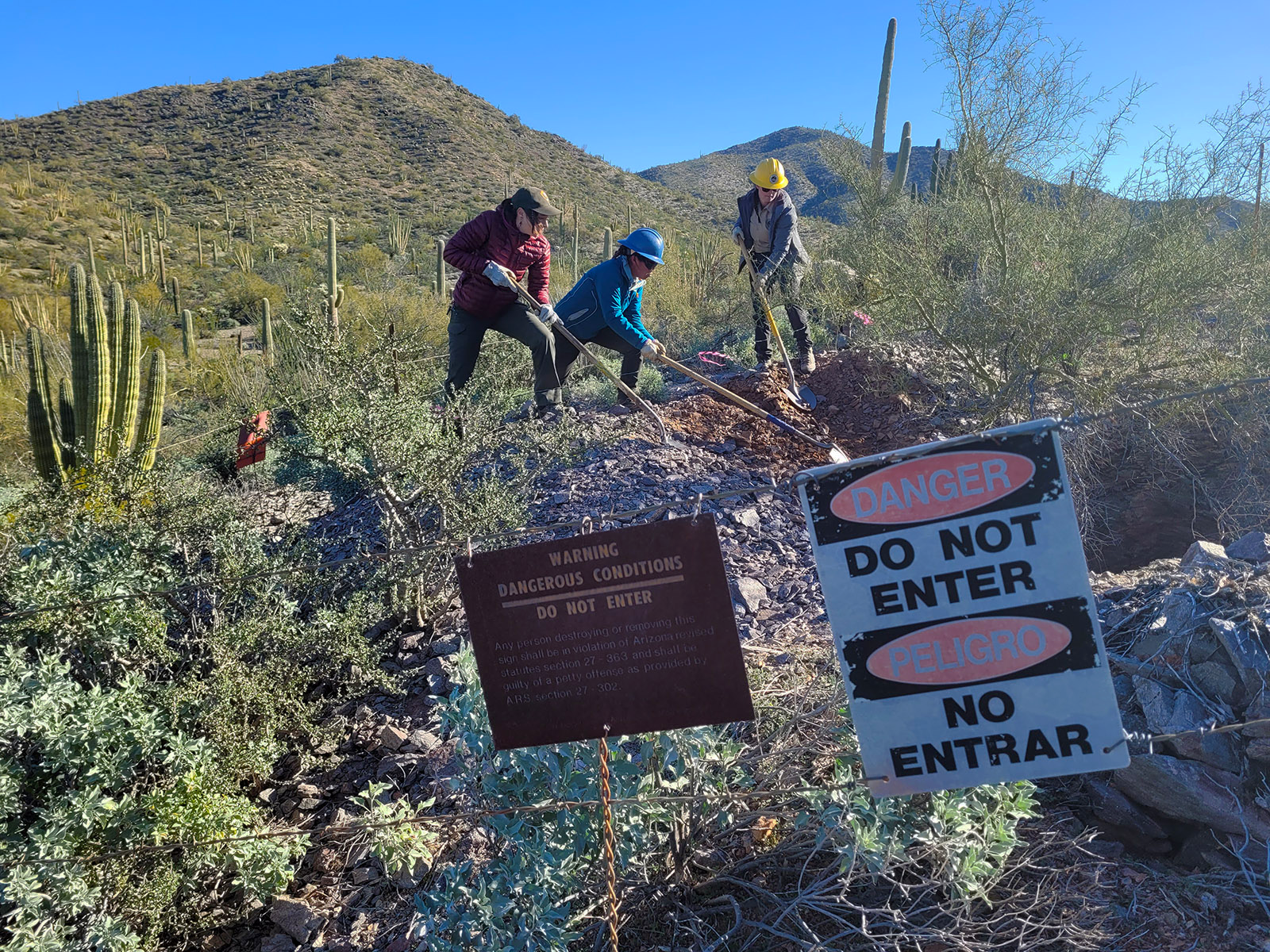 three people stand behind a barbed wire fence with danger signs and use shovels to fill in an abandoned mine