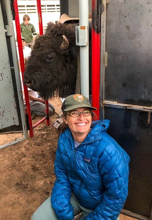 In the foreground, a woman wearing a blue jacket is smiling as a bison's head is looking through a doorway.