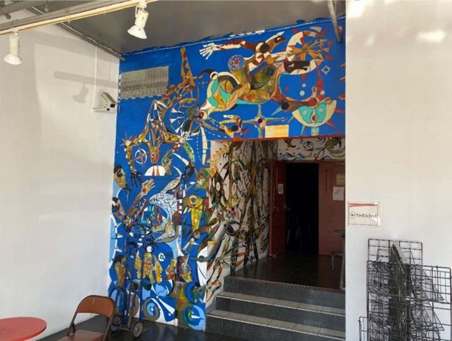 Narrow portion of wall over and adjoining doorway covered from floor to ceiling in blue background with mural of yellow-green and brown geometric animal figures