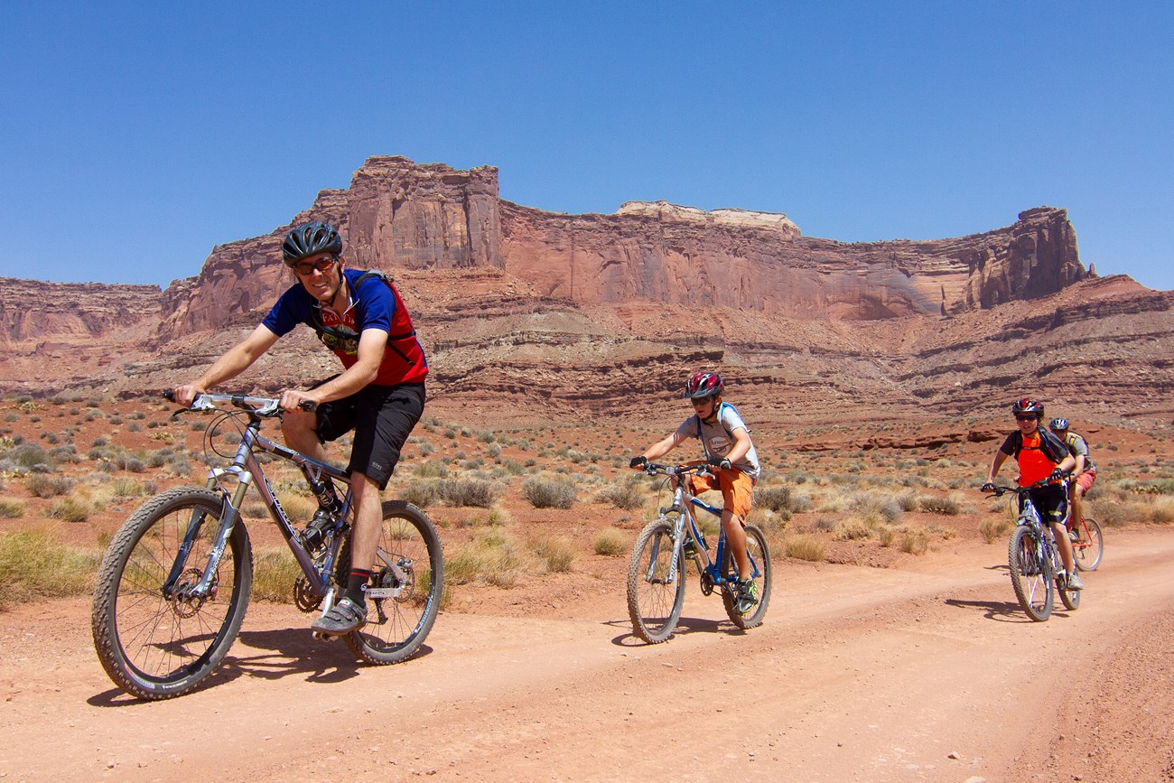 four mountain bikers with helmets and packs bike down a dirt trail in the desert