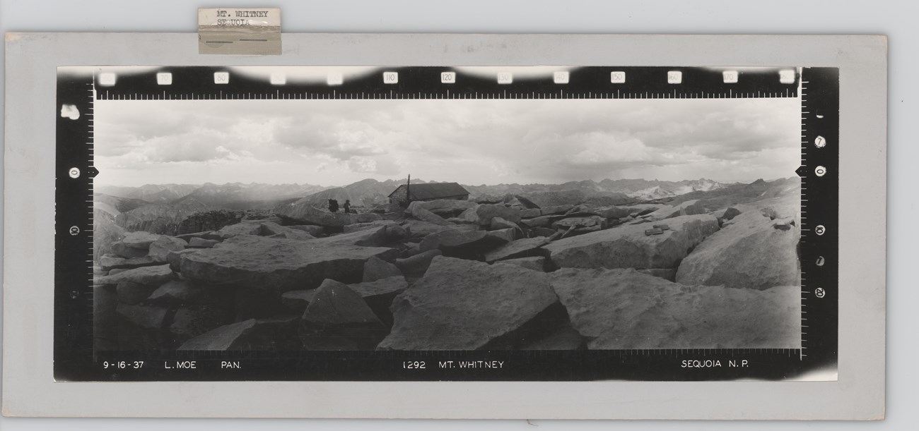 Mount Whitney, Sequoia Panoramic photograph of a large boulder field with a small building in the background. A man and pack animal are outside the building.
