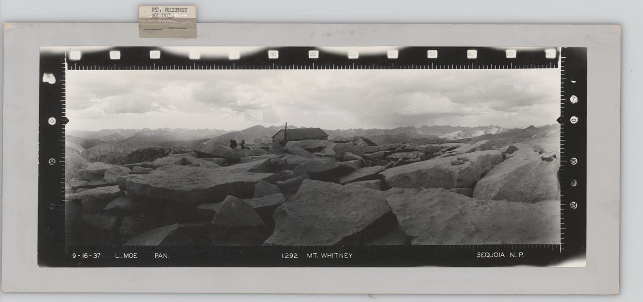 Mount Whitney, Sequoia Panoramic photograph of a large boulder field with a small building in the background. A man and pack animal are outside the building.