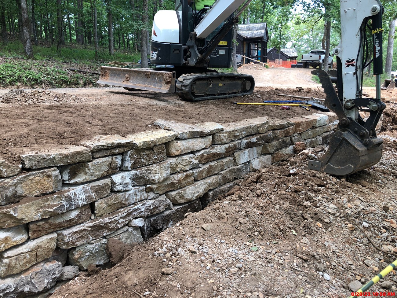 Cabin 404 Retaining Wall A construction site in the woods with a stone retaining wall that is being worked on with cabins in the background.