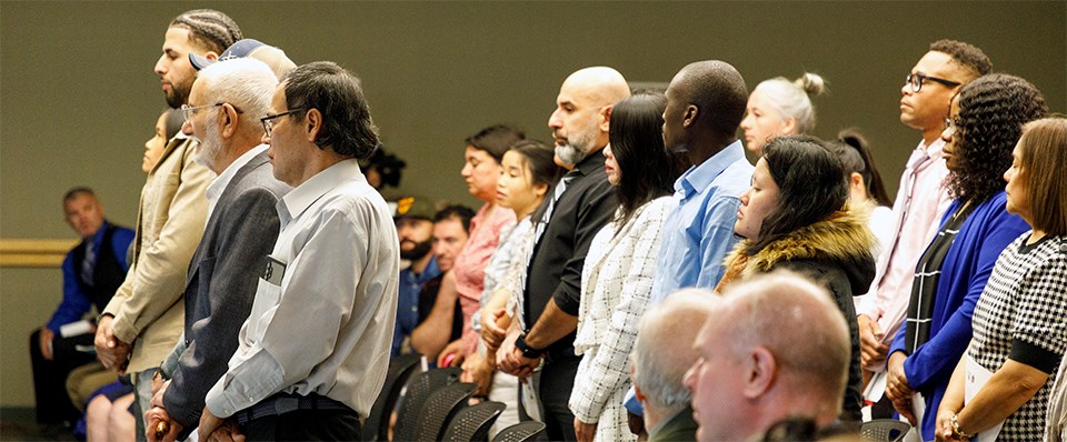 New citizens prior to taking the oath of citizenship. A group of men and women of many nationalities stand facing the front of a room.