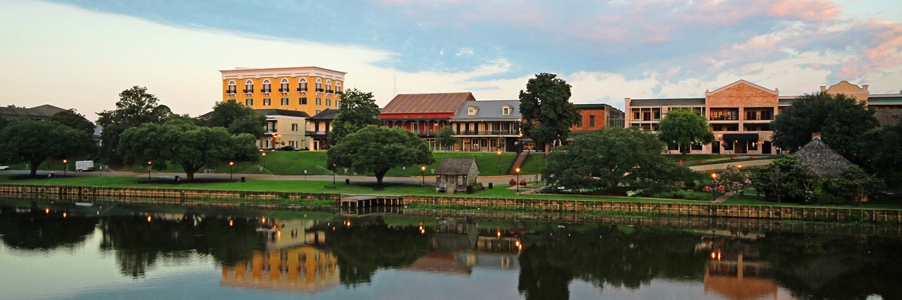 Riverfront along Front Street in Natchitoches, Louisiana, taken from the opposite bank. Two story buildings dominate the roofline, with a bank and apartments rising to four and three stories, respectively.