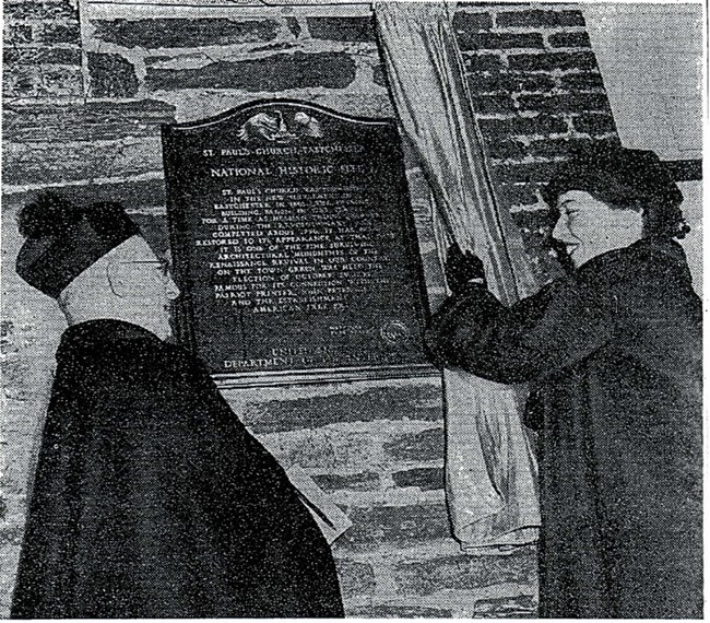 Man in priestly dress and a woman standing in front of a metal plaque, attached to a stone wall