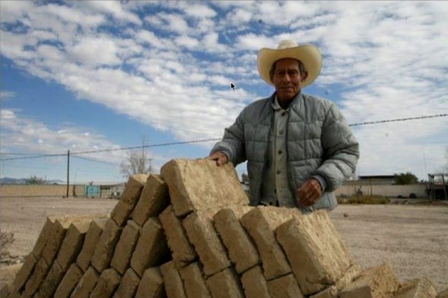 Abodero elder standing with adobe blocks, construction materials.