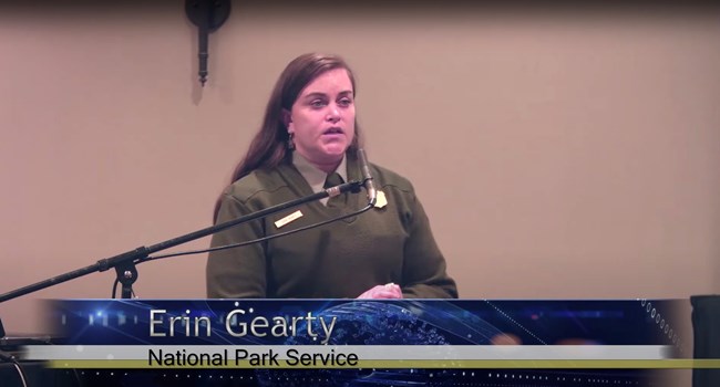 The speaker Erin Gearty stands at a lectern. Watch a non-audio described version of this presentation on YouTube.