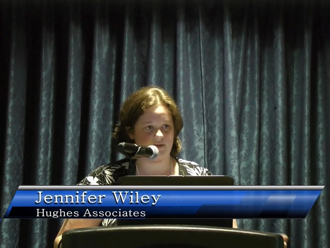 Still photo of speaker Jennifer Wiley presenting from a laptop at a lectern.