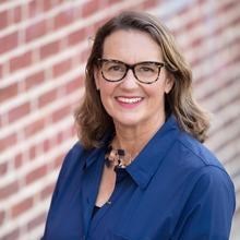 Anglo woman with spectacles and shoulder length hair in a blue blouse against a masonry backdrop.