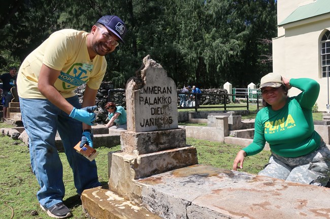 A man and woman smile as they clean a concrete grave marker.