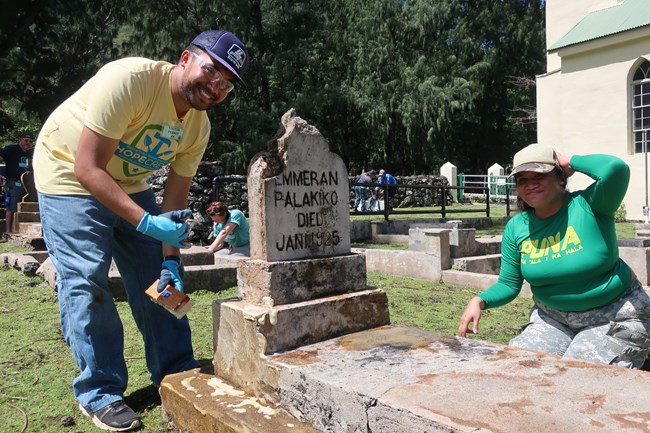 Hilo students scrub a concrete grave marker removing biological growth. A man and woman smile as they clean a concrete grave marker.