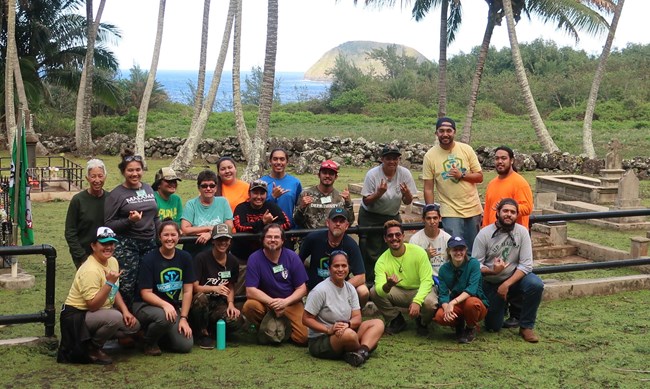 Recent HOPE Crew at Kalaupapa National Historical Park. About two dozen people beside a cemetery with palm trees and the ocean in the background.
