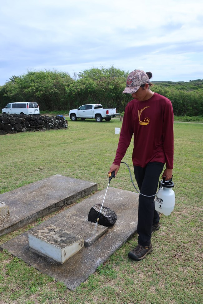 Cody Koʻokāne Pacheco applying D/2 to a grave before scrubbing with a natural bristle brush A slender man spraying a grave and marker with D/2.