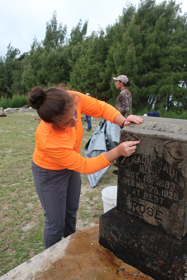 Student uses a bamboo skewer to remove biological build up from the inscription. Student uses a bamboo skewer to remove biological build up from the grave marker inscription.