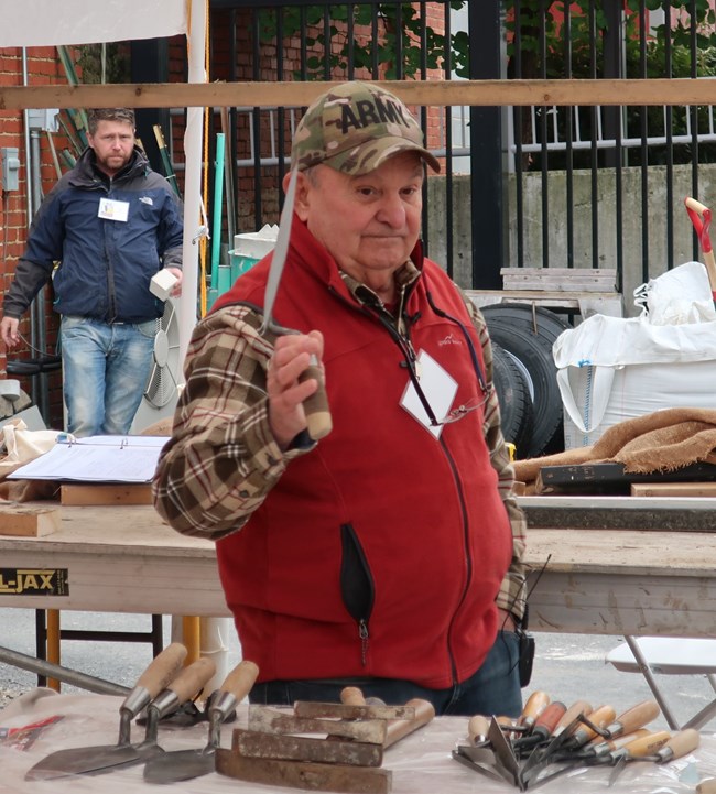 Dom DeRubis holding up a trowel and standing in front of a set of masonry tools.