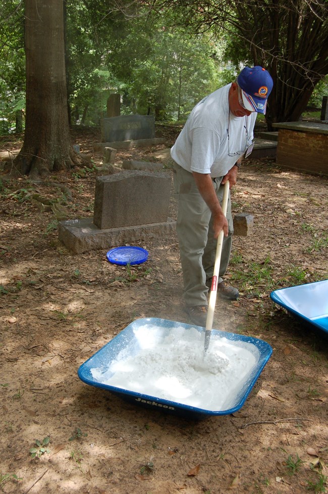 Dom DuRubis mixing quick lime with a shovel in a shallow container within a cemetery.