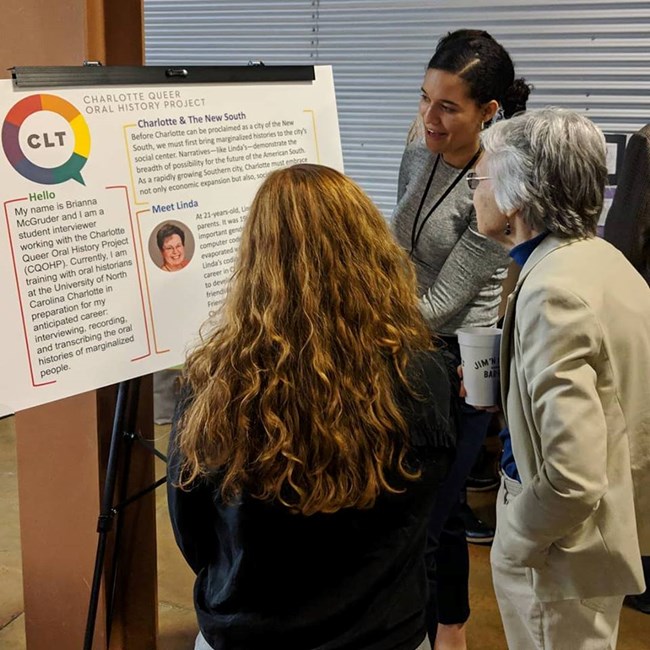 Poster presentation on the Charlotte Queer Oral History Project Three women gather around a poster.