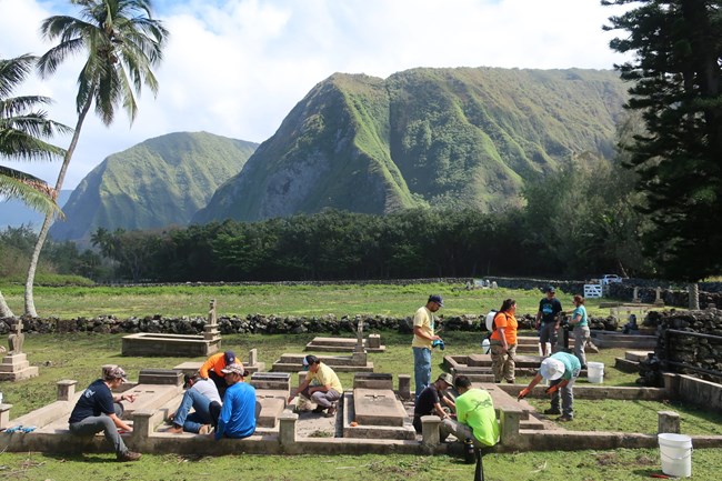 A crew of people cleaning graves at the Kalawao Cemetery with mountains in the background.