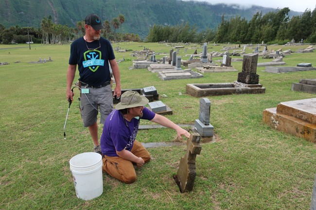 Rusty Brenner (standing) and Jason Church discuss a marker during cleaning within a cemetery with mountains in the background.
