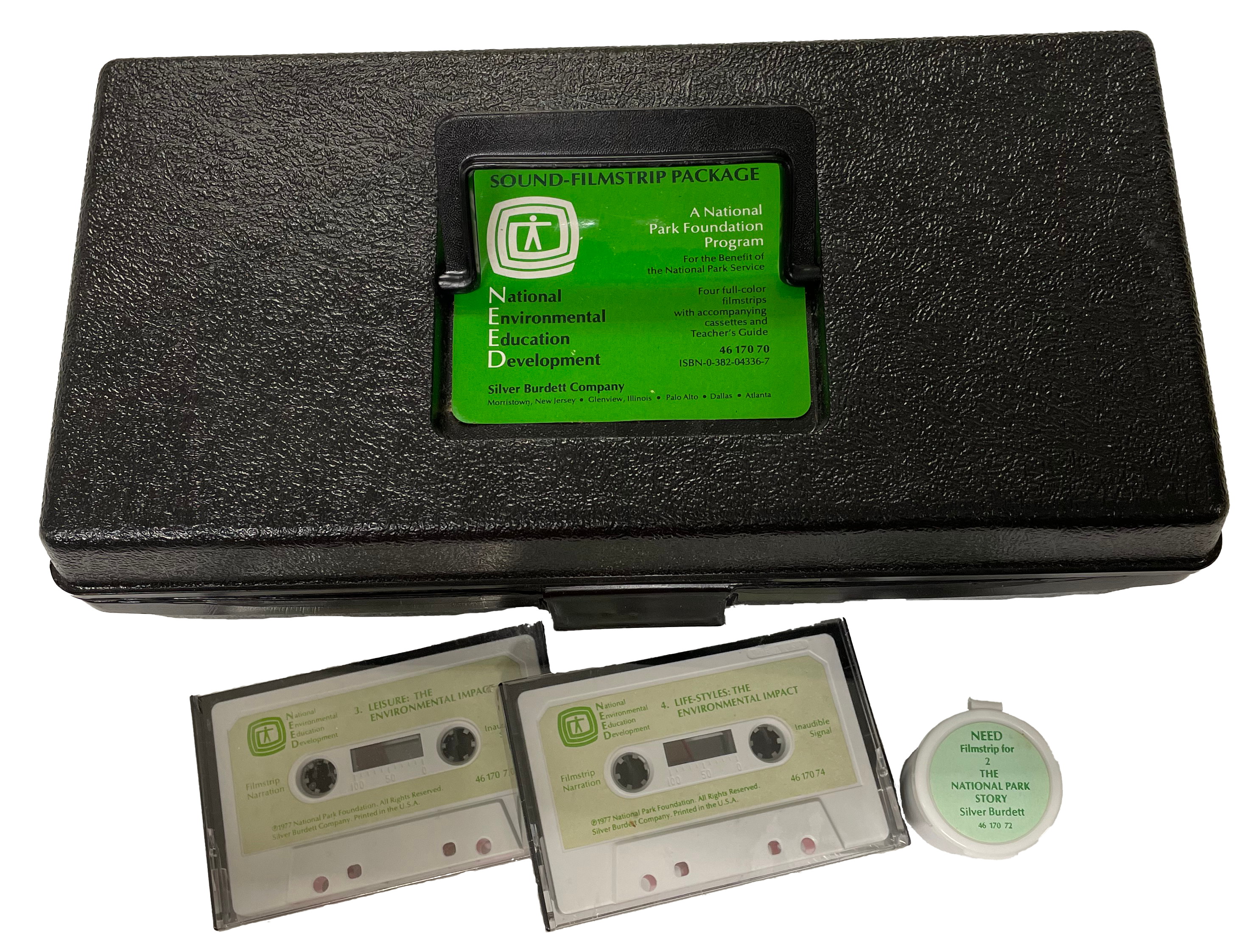 Plack plastic box with green label. Two audiocassettes and a filmstrip cannister beside it.