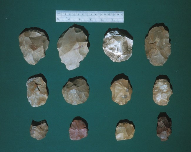 lt= twelve small (around 1 inch in length) worked stones sit on a green fabric with a scale bar.
