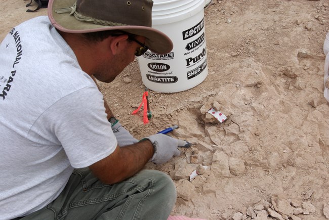 a man looks down at various tools like a hammer and pick, as well as the hard sandstone they sit on.