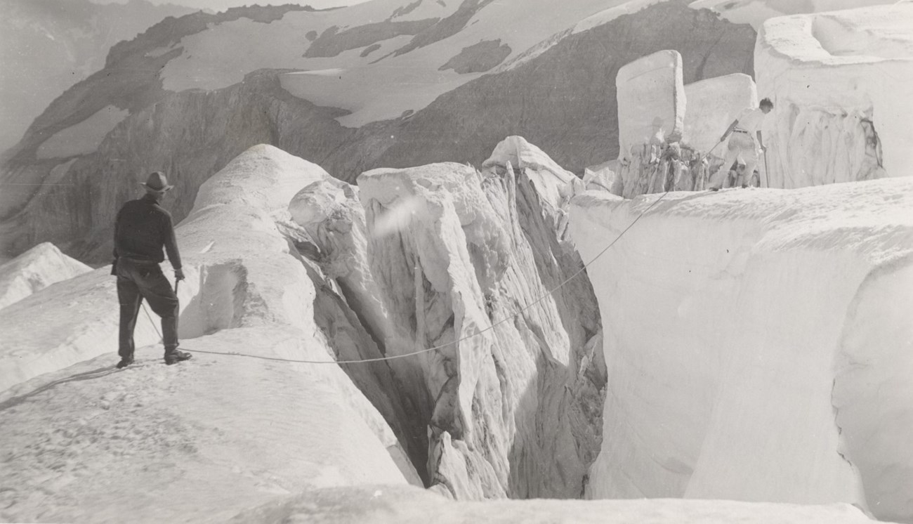 Two men stand on opposite sides of a crevasse with a rope across it.