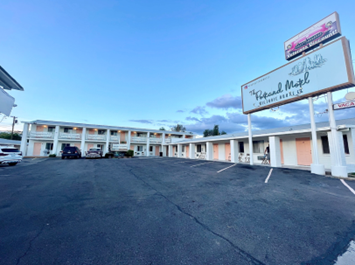 lt= from the parking lot, a one-story, white colored motel building with a wraparound veranda.
