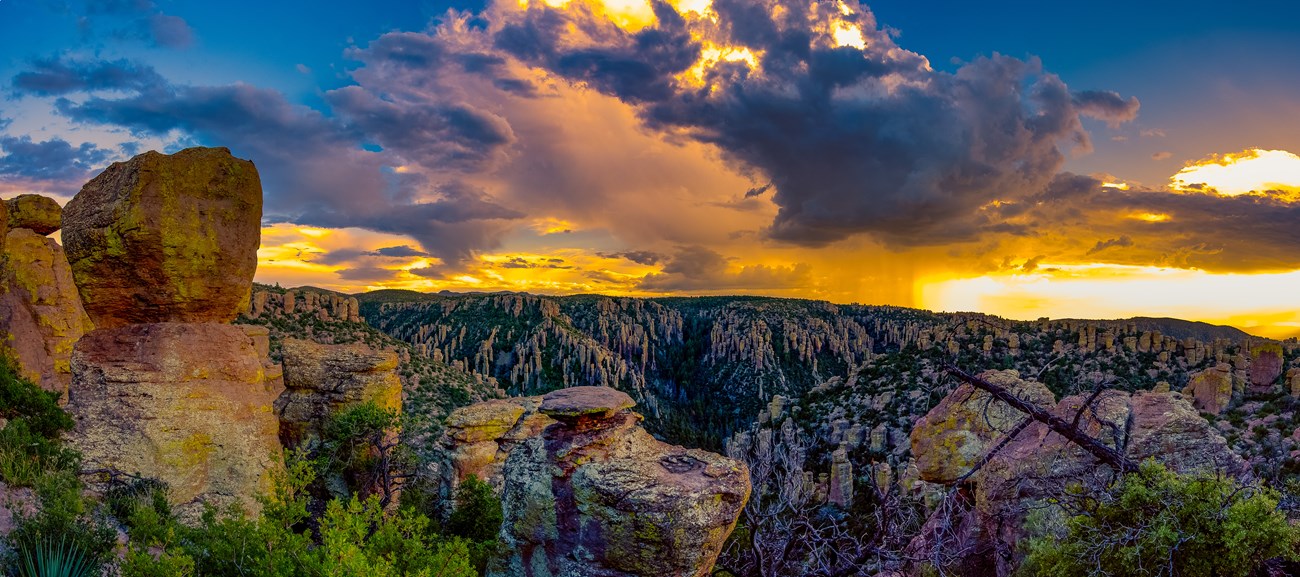 The sun sets behind monsoon clouds and rain, illuminating hoodoos in bright orange and blue.