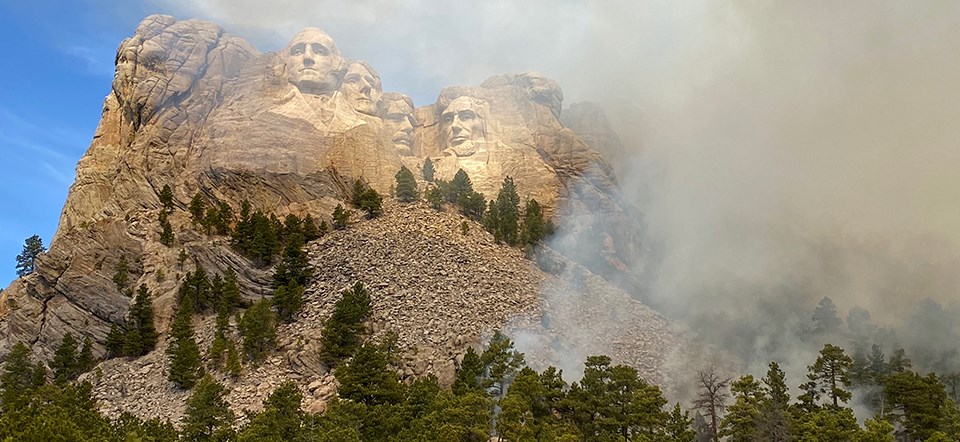 Smoke rises from trees on slope below carved figures on Mount Rushmore, obscuring view.