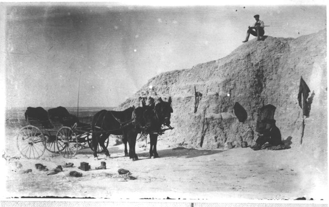 a historic photo shows a wooden horse-drawn wagon next to a butte with a man sitting on top holding a rock hammer.
