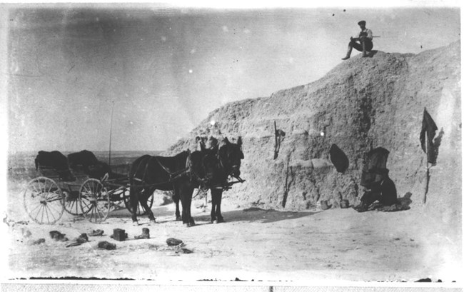 lt= a historic photo shows a wooden horse-drawn wagon next to a butte with a man sitting on top holding a rock hammer.