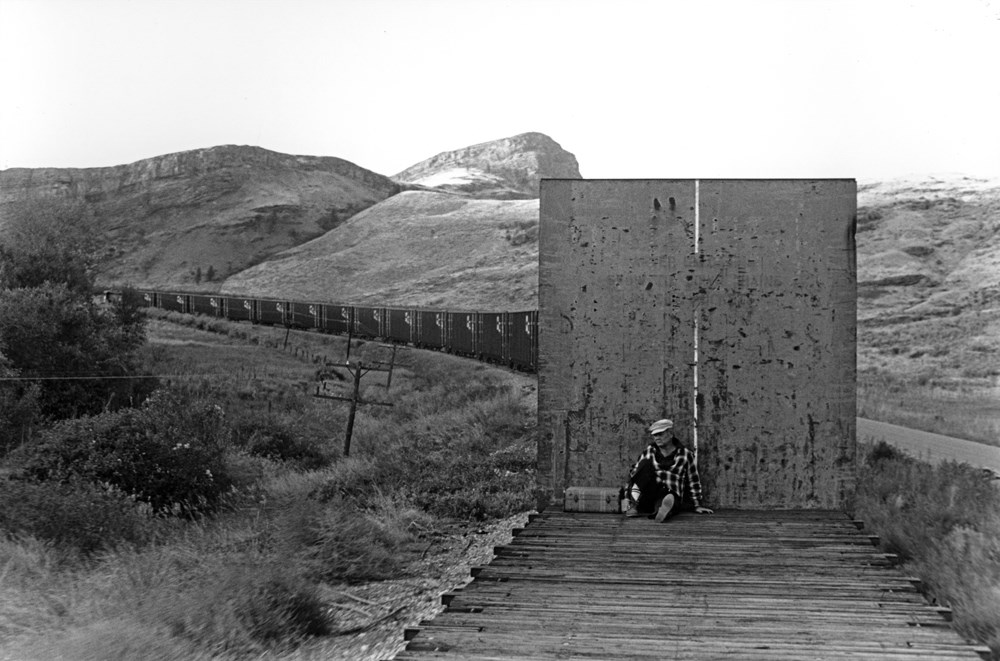 Black and white photo of man on a flat train car in the mountains