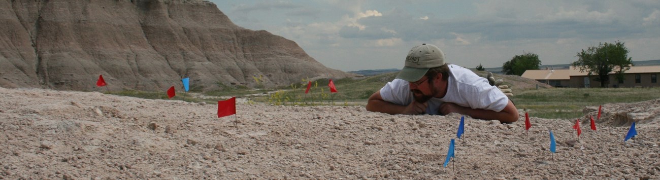 a man lies on the ground among different color flags with rugged badlands formations and a squat building in the background.