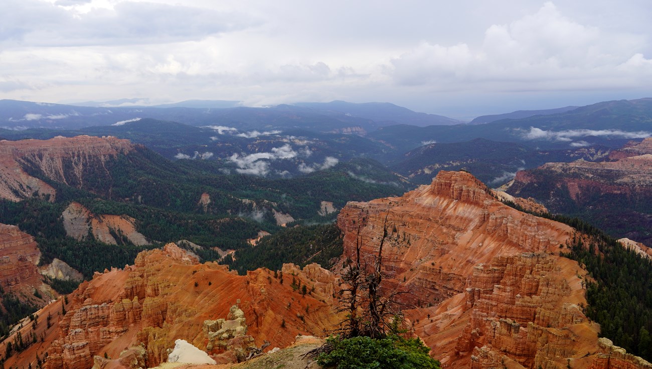 This scenic view from Cedar Breaks shows dramatic red cliffs, forested slopes, and drifting clouds. Layered red cliffs descend into forested canyons beneath drifting mist and clouded sky.