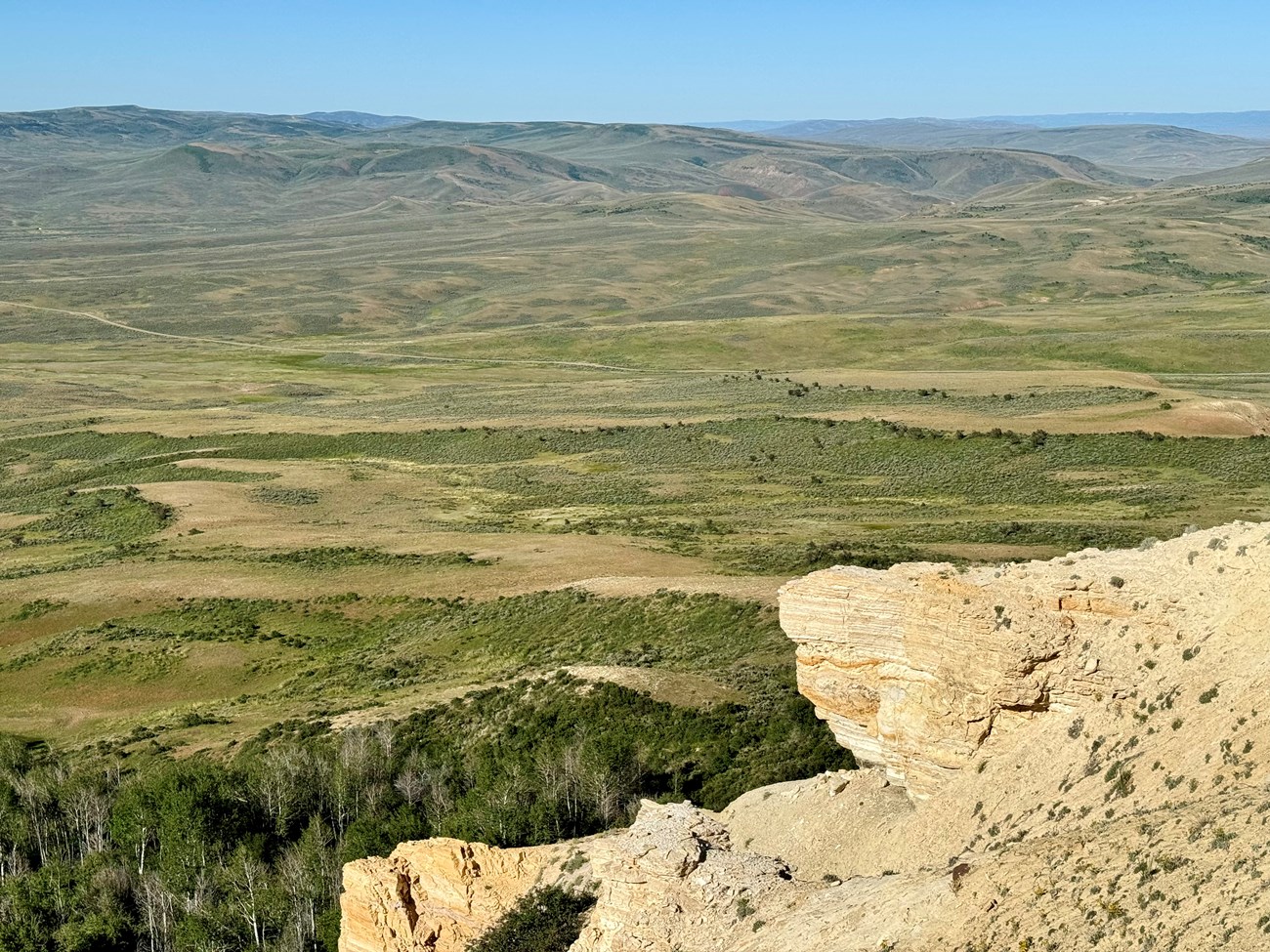 panoramic-view-fossil-butte-national-monument Panoramic view from a high point at Fossil Butte National Monument showing sagebrush-covered lowlands, scattered green tree stands, and a prominent tan sandstone outcrop in the foreground.