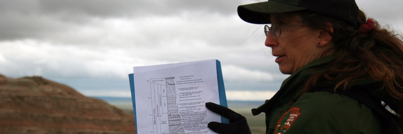 a women with a ponytail and NPS baseball hat points at a geologic diagram while standing in front of badlands formations.