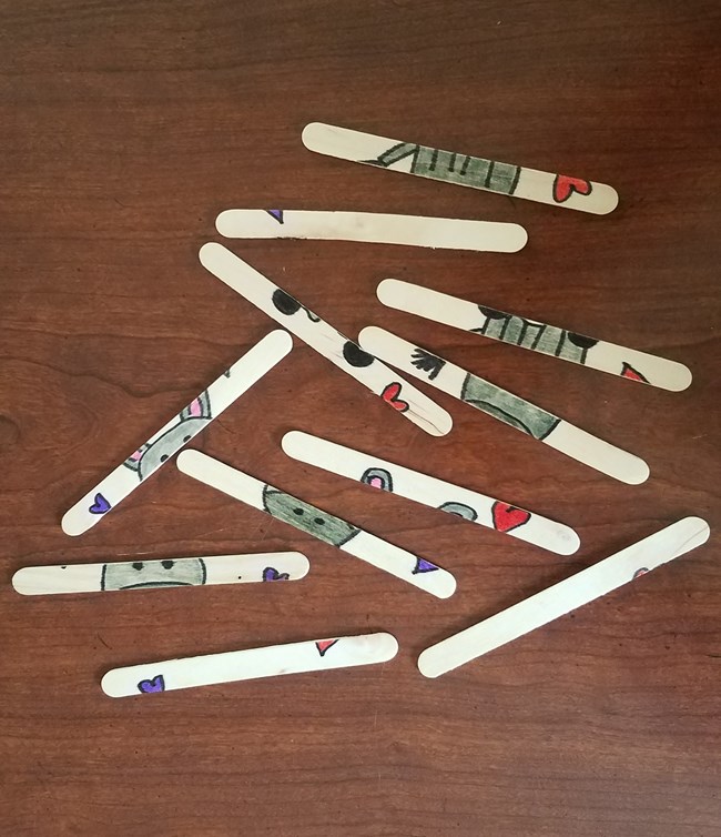 Craft sticks spread out on a wooden table; each contains part of a drawing made mostly with gray marker.