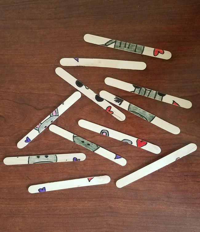 past people craft Craft sticks spread out on a wooden table; each contains part of a drawing made mostly with gray marker.