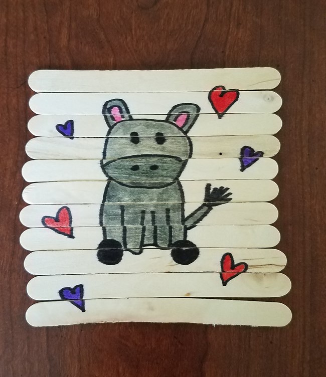 Craft sticks lined up to reveal a drawing of a grey hippo surrounded by red and purple hearts.