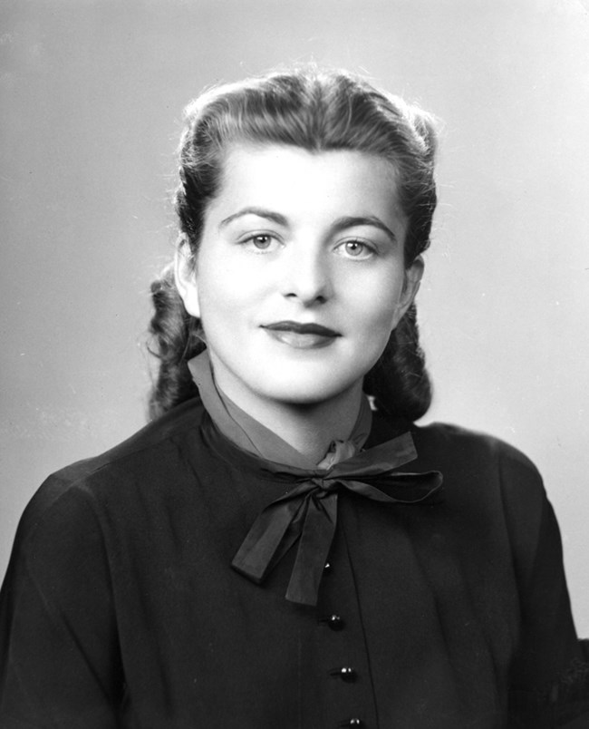 A black and white photo portrait of a young woman with light eyes and light wavy hair styled back in rolls, wearing a dark long sleeved bow blouse and lipstick, looking to the viewer and smiling.