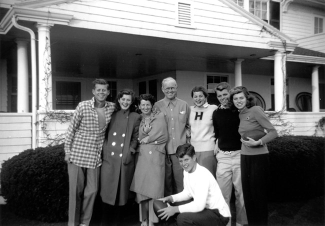 A black and white photo of the Kennedy family in casual attire.  They stand in front of a white house.  Ted Kennedy kneels in the front with a football.
