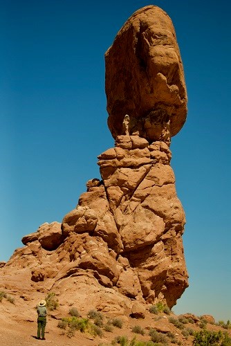 a ranger gazes up at a large rock balanced on a sandstone pillar
